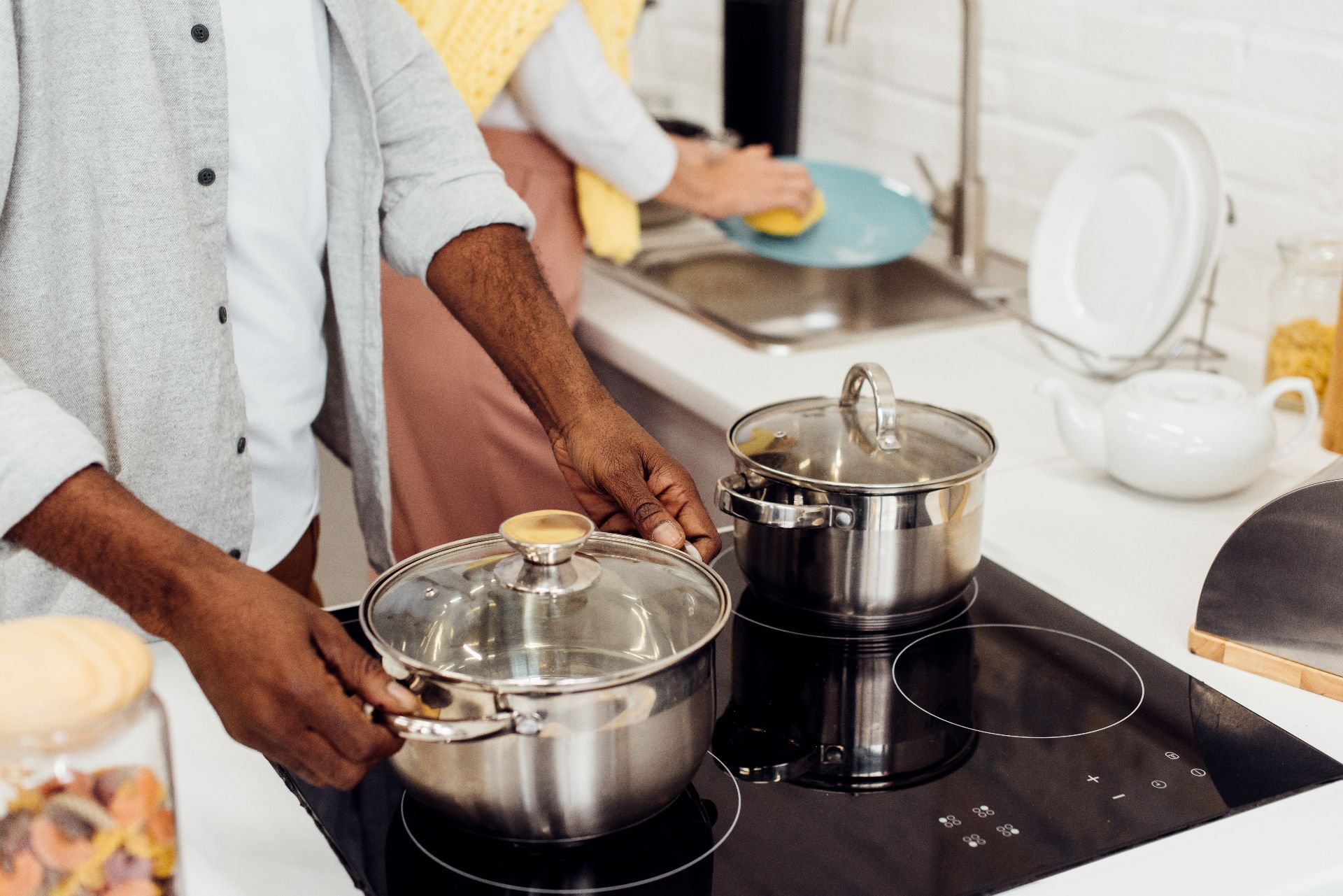 Man handles stainless steel pot on stove as woman washes dishes in the background - All-Clad stainless steel