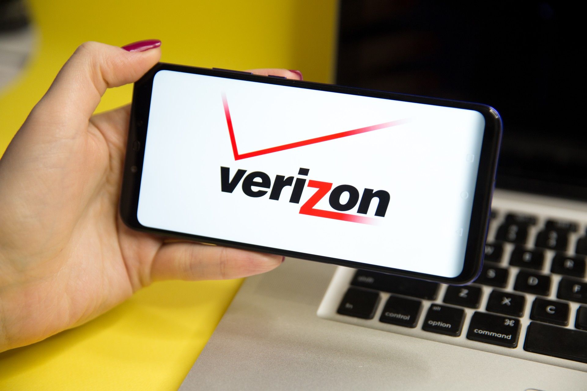 A woman's hand holds a cellphone with a Verizon screen in front of a laptop - Verizon bill