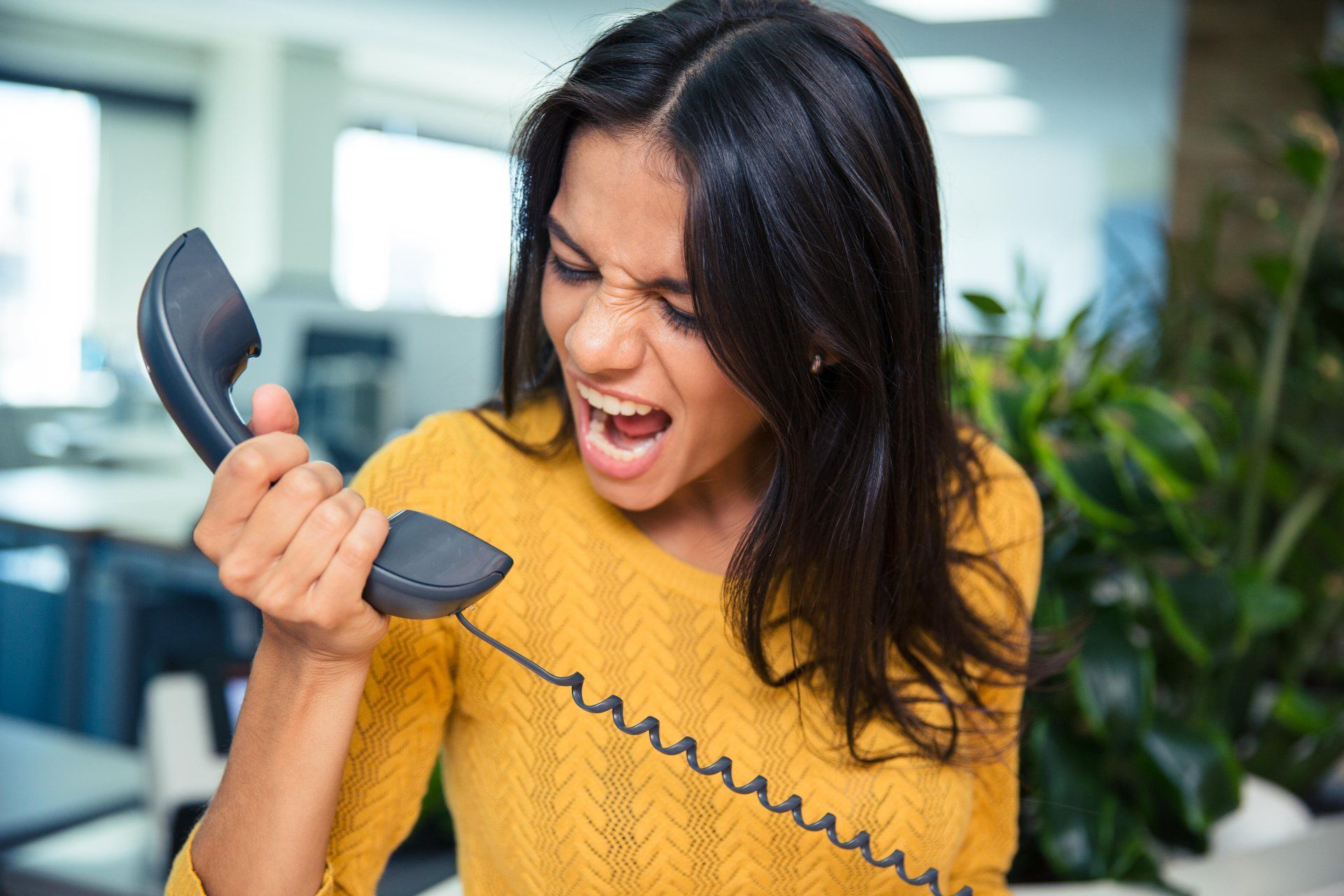 A dark-haired woman in a yellow top yells into a phone receiver she is holding away from her face - telephone calls