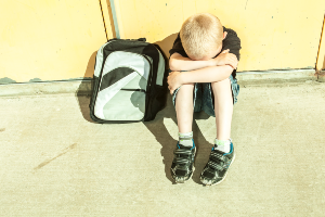 A sad boy sits next to a backpack, hiding his face in his arms on top of his knees pulled to his chest - ymca