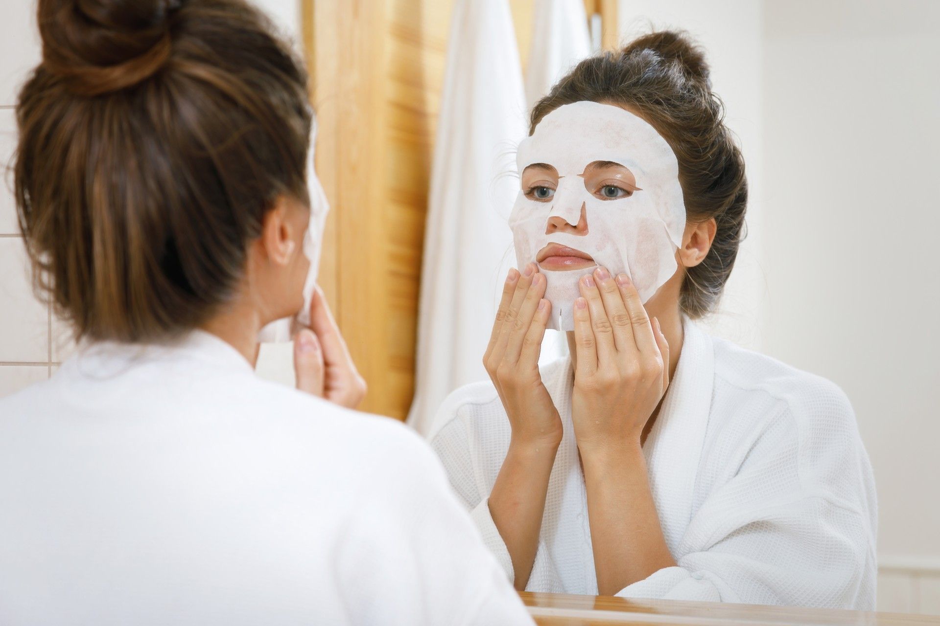 A woman in a white robe looks in a mirror as she puts on a sheet mask - Yes to Grapefruit