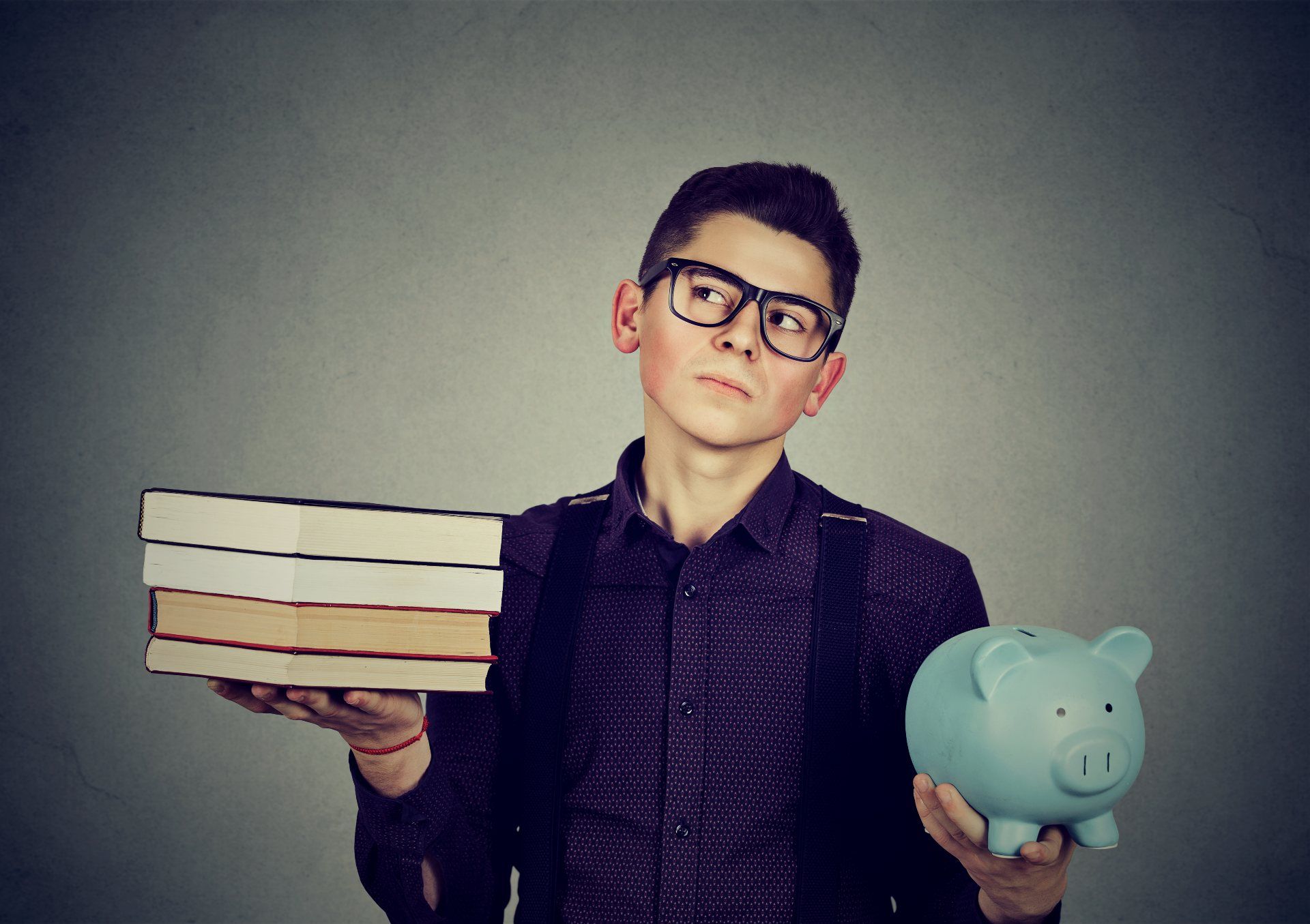 A young man wearing glasses and a backpack holds a stack of four books in his right hand and a blue piggy bank in his left hand - pheaa