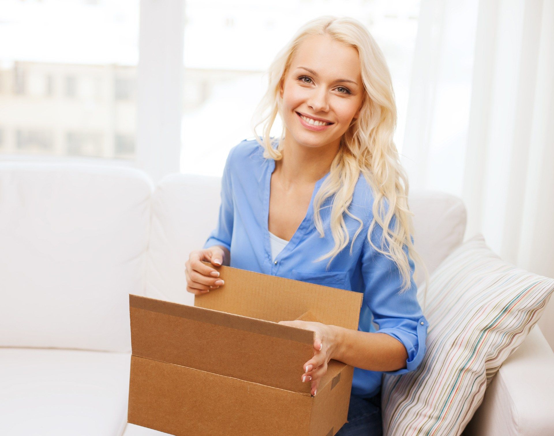 A smiling blonde woman opens a cardboard box while sitting on a sofa - fabfitfun