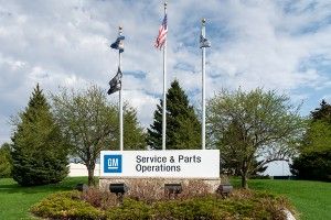 A sign reads "GM Service & Parts Operations", with four flags on three poles behind it