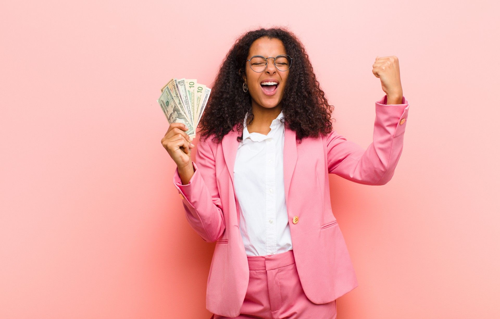 A woman in a pink suit standing against a rosy background closes her eyes and holds up a handful of fanned-out cash while pumping her fist with the other hand.