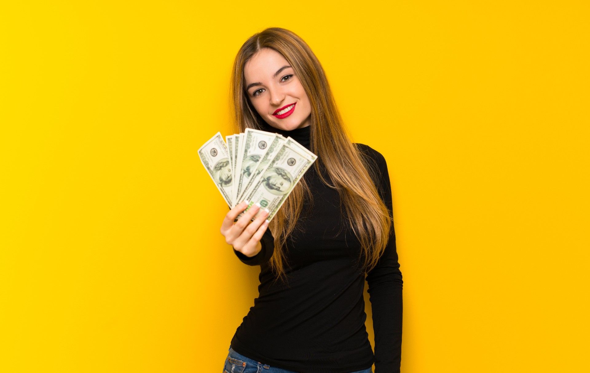 A long-haired woman in a black turtleneck holds fanned-out cash up to the camera while standing against a yellow background