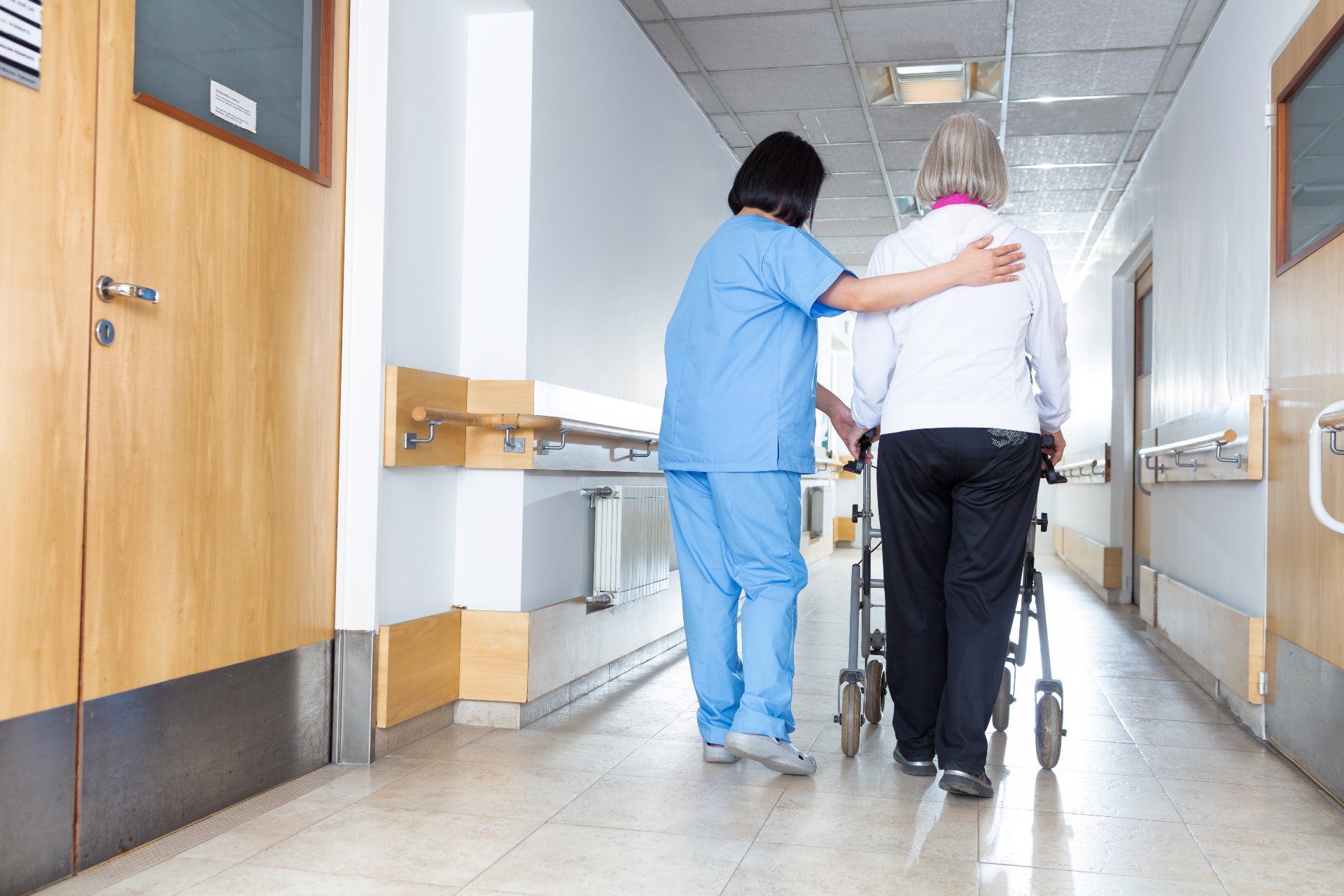 A nurse in blue scrubs helps a patient walk down a hallway at a nursing facility or hospital - Prompt Nursing