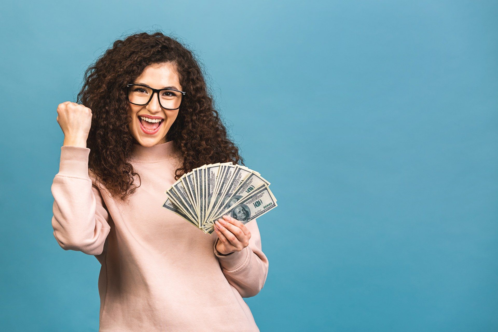 A happy woman wearing glasses celebrates while holding cash against a blue-green background