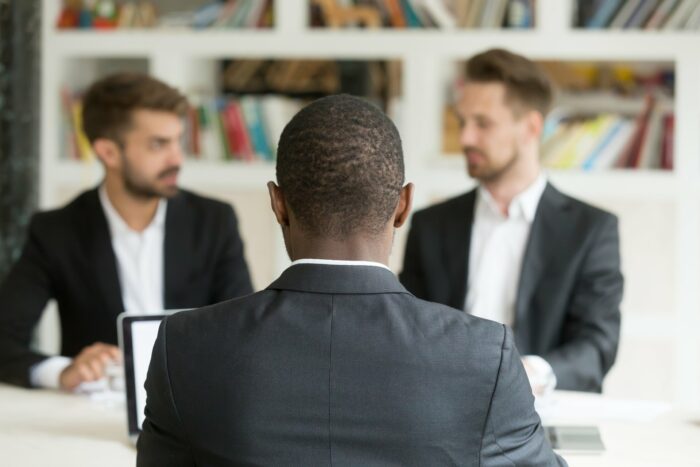 Black male employee in suit sits across from two caucasian male employees - Personnel Staffing Group - Race discrimination - mvp