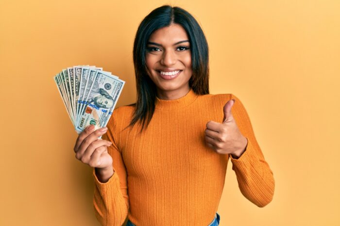 A smiling woman in an orange sweater holds up fanned out cash and gives a thumbs up