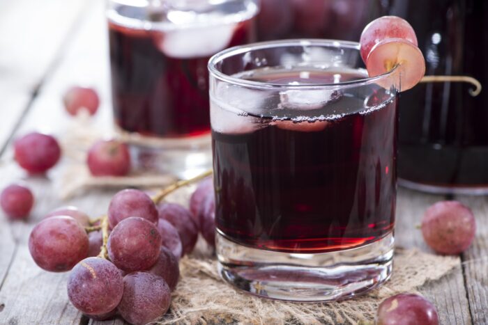 Purple grape juice in a glass with ice, surrounded by grapes with a grape garnish on the rim of the glass - welch's juice