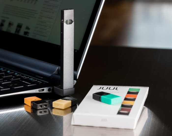 Juul e-cigarette or nicotine vapor dispenser being charged with box and JUULpods on table