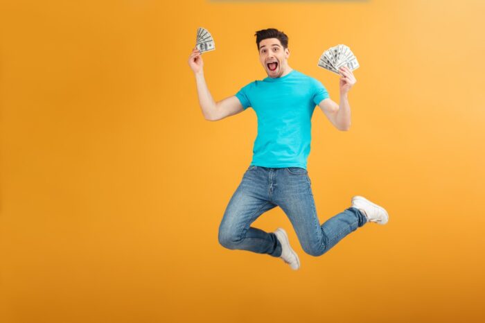 Portrait of a joyful young man in t-shirt holding bunch of money banknotes and celebrating while jumping isolated over yellow background.