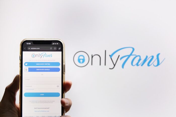 View of a hand holding up a smartphone displaying the OnlyFans logo on the screen against an OnlyFans logo in the background, representing the Facebook and OnlyFans class action lawsuit.