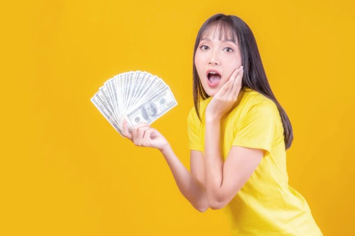 Excited beautiful Asian business young woman cute girl with bangs hair style in yellow t shirt holding money US dollar bills in hand
