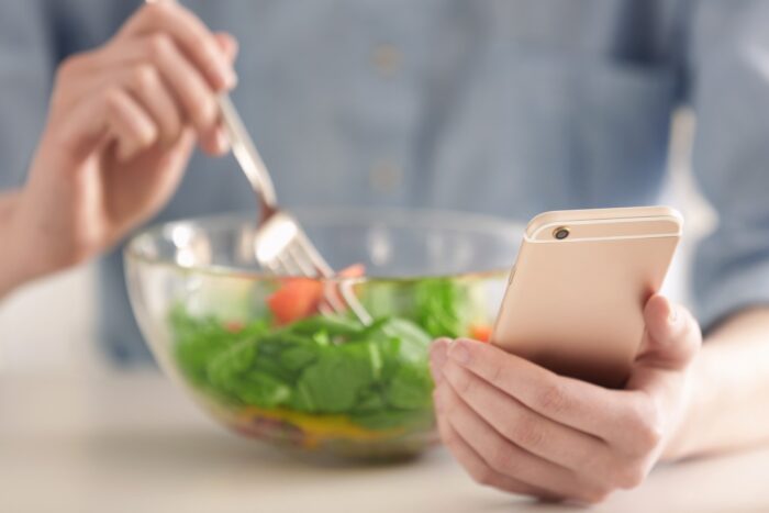 A woman uses a fork to eat salad from a glass bowl while holding a smartphone in her other hand - noom settlement