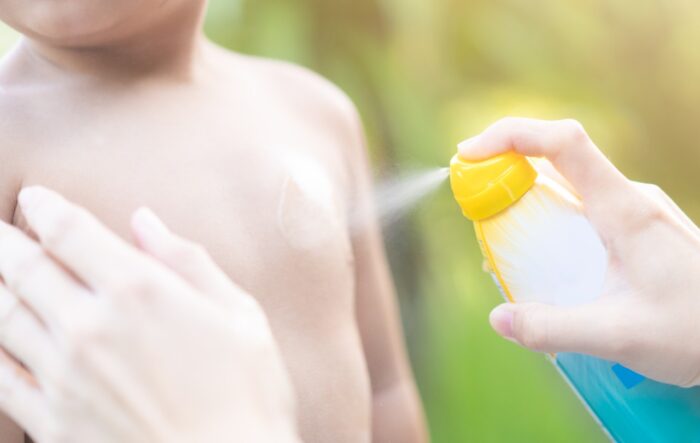 Mother applying sun screen spray on her baby boy in at the sea beach for swim - sunscreen carcinogens - neutrogena class action lawsuit - sun product settlement