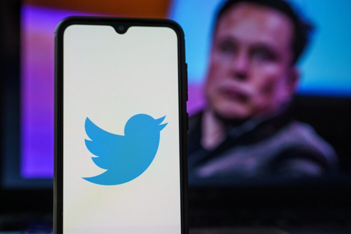 Photo of Twitter logo on a smartphone with Elon Musk in the background.