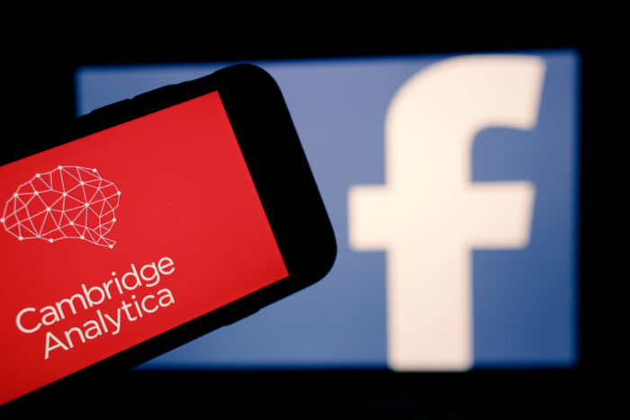 The logo of the strategic communication company Cambridge Analytica is seen on the screen of an iPhone in front of a computer screen showing Facebook logo.