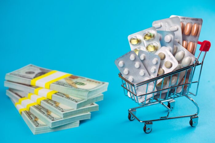 High generic drug prices concept. shopping cart filled red medicinal capsules tablets pills, wad of dollar money, blue background - nexium settlement, diovan class action lawsuit