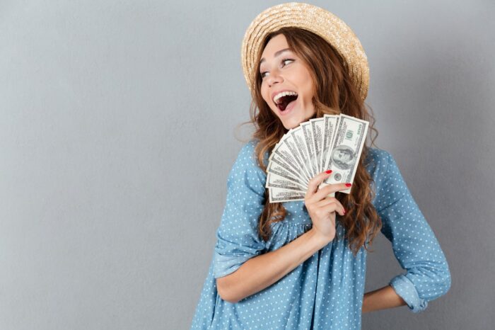 Picture of excited young happy woman standing over grey wall wearing hat holding money.