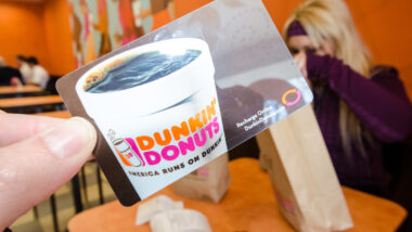 A hand holds up a Dunkin Donuts gift card inside of a Dunkin Donuts franchise store eating area.