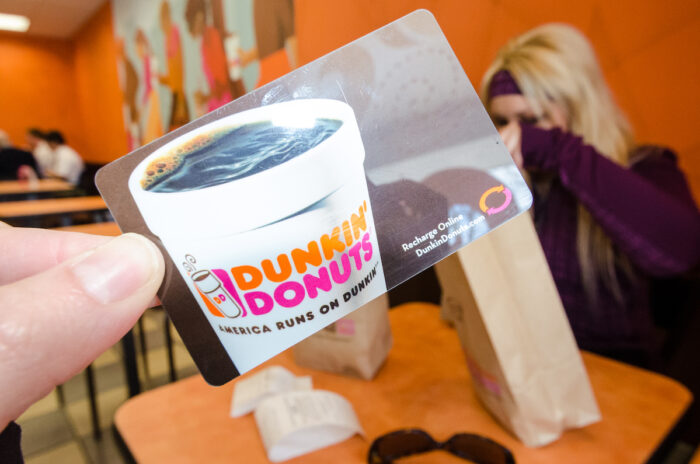 A hand holds up a Dunkin Donuts gift card inside of a Dunkin Donuts franchise store eating area, representing the Dunkin' non-refundable gift cards class action.