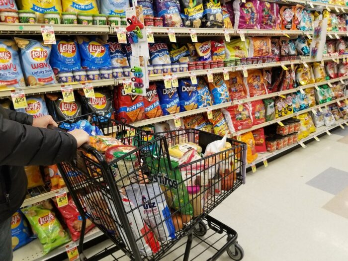 Pushing a cart filled with groceries on the snacks aisle inside a supermarket.