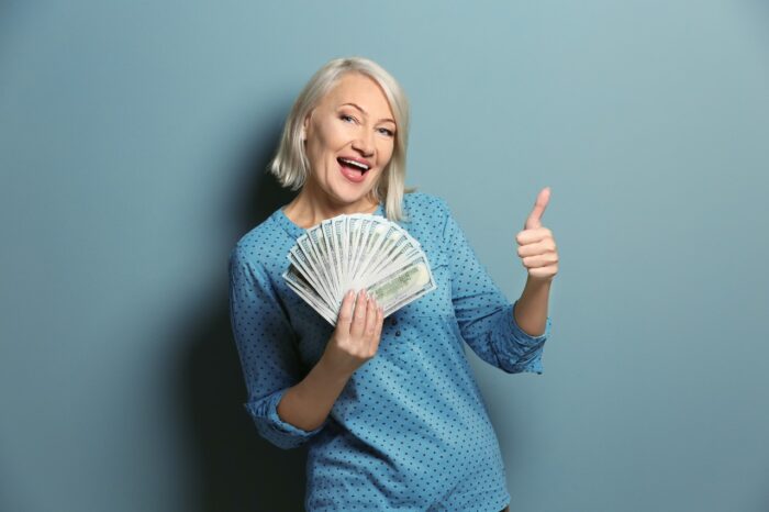 Happy mature woman with money showing thumb-up gesture on color background, settlement checks
