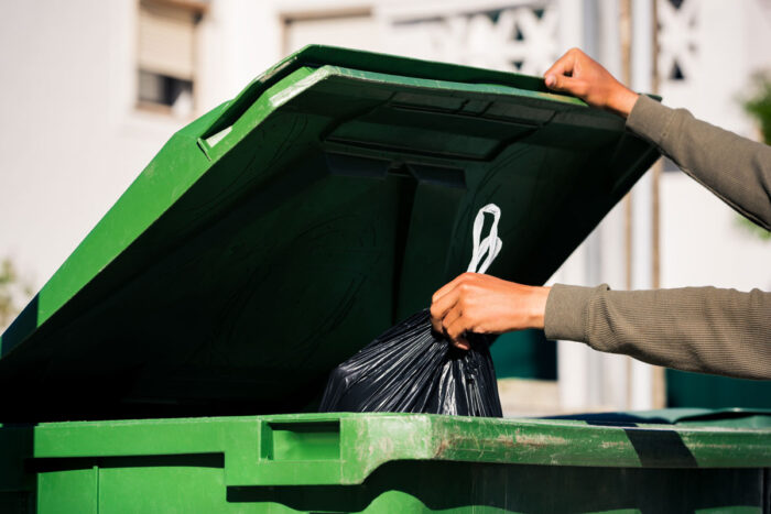 Man throwing out black eco-friendly recyclable trash bag in to big plastic green garbage container - waste pro environmental fees class action lawsuit
