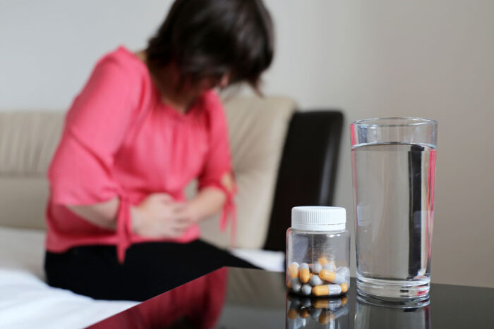 Bottle of pills and water glass on a table, girl sitting on bed clutching her belly.