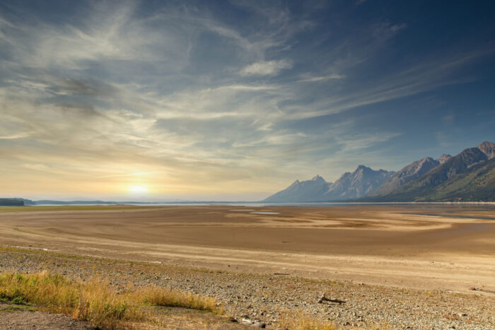 Jackson Lake drought causing receding water and dry lake bed due to global warming at sunrise or sunset in Grand Teton National Park with mountains in background.