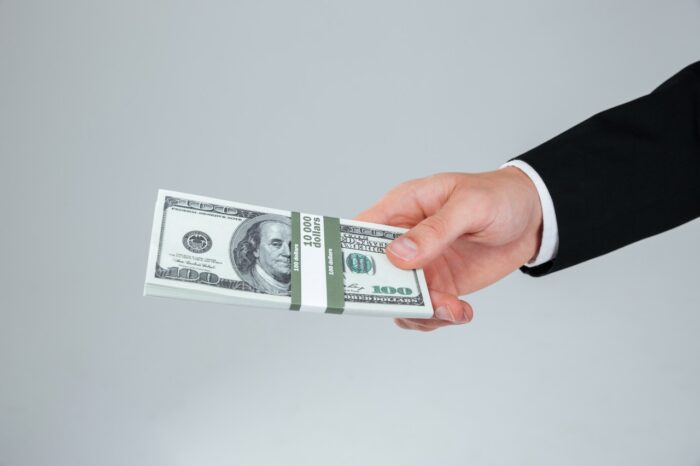 Hand of businessman in suit holding money over grey background, FTC fraud refund concept.
