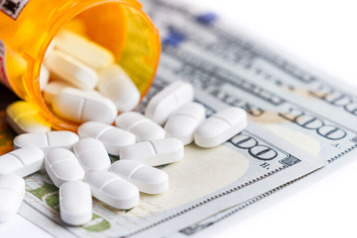 Close up of a prescription bottle with white tables falling out on top of a group of hundred dollar bills.