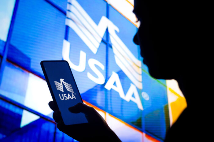 In this photo illustration, a woman's silhouette holds a smartphone with the United Services Automobile Association (USAA) logo displayed on the screen and in the background - usaa class action lawsuit, settlement, total loss