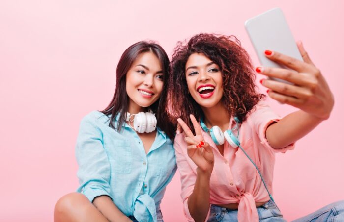 Two women with headphones around their neck take a selfie with a smartphone in front of a pink background - snapchat settlement