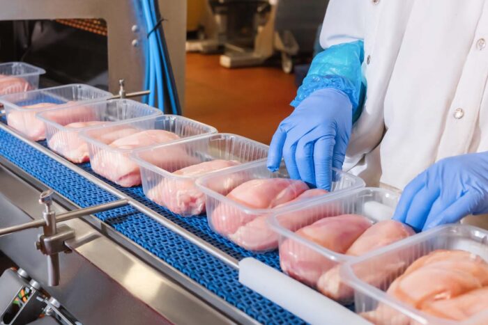 Close up of raw chicken on a conveyer belt being packaged by a factory worker.