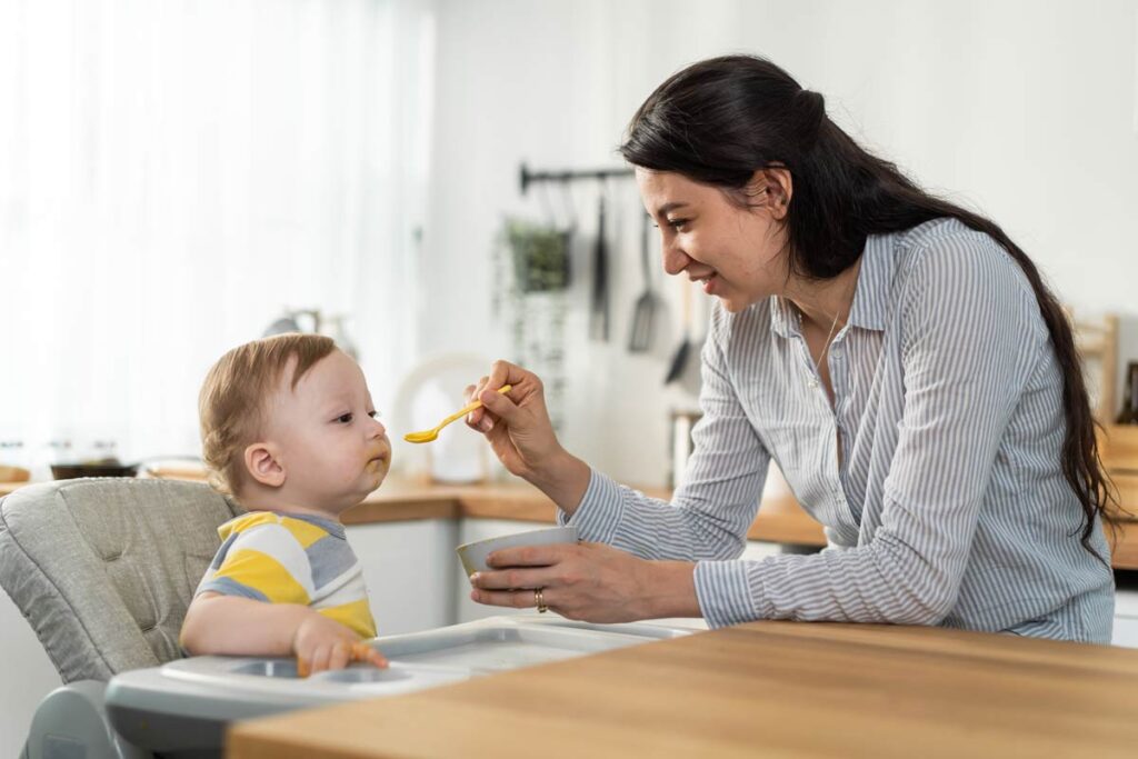A mother giving her baby food on a spoon inside a kitchen.