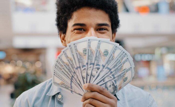 Happy wealthy successful smiling african american business man looking at camera holding fan of money hiding behind dollar banknotes.