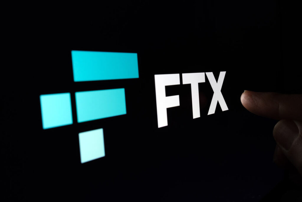 Finger pointing at blurred FTX Cryptocurrency Exchange company logo on dark display.