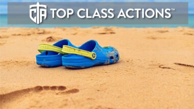 A pair of blue with a yellow strap Crocs sandals are on a sandy beach.