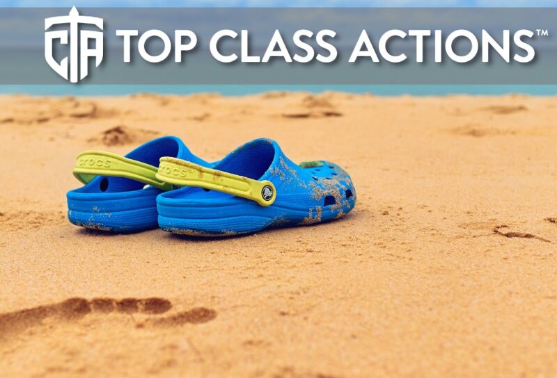 A pair of blue with a yellow strap Crocs sandals are on a sandy beach.