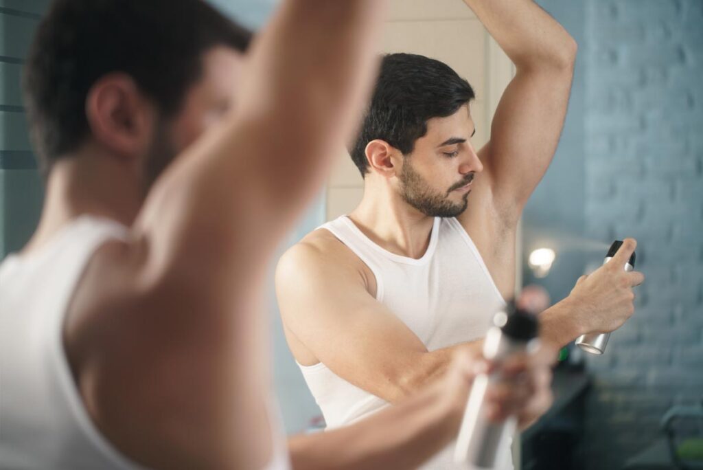 Confident man using spray deodorant on underarm skin, smiling and looking at mirror, representing the Brut and Sure antiperspirant benzene class action lawsuit settlement.