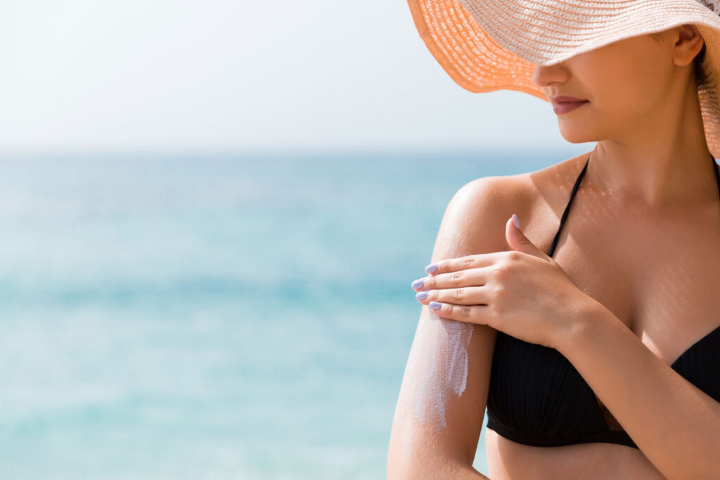 Woman in a hat putting sunscreen on shoulder outdoors under sunshine on beautiful summer day.