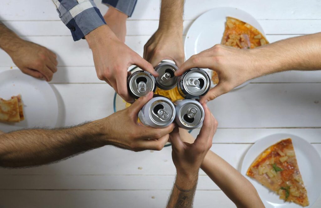 Top view group of best friends sitting at the table and clicking aluminum cans with soda on wooden table background.