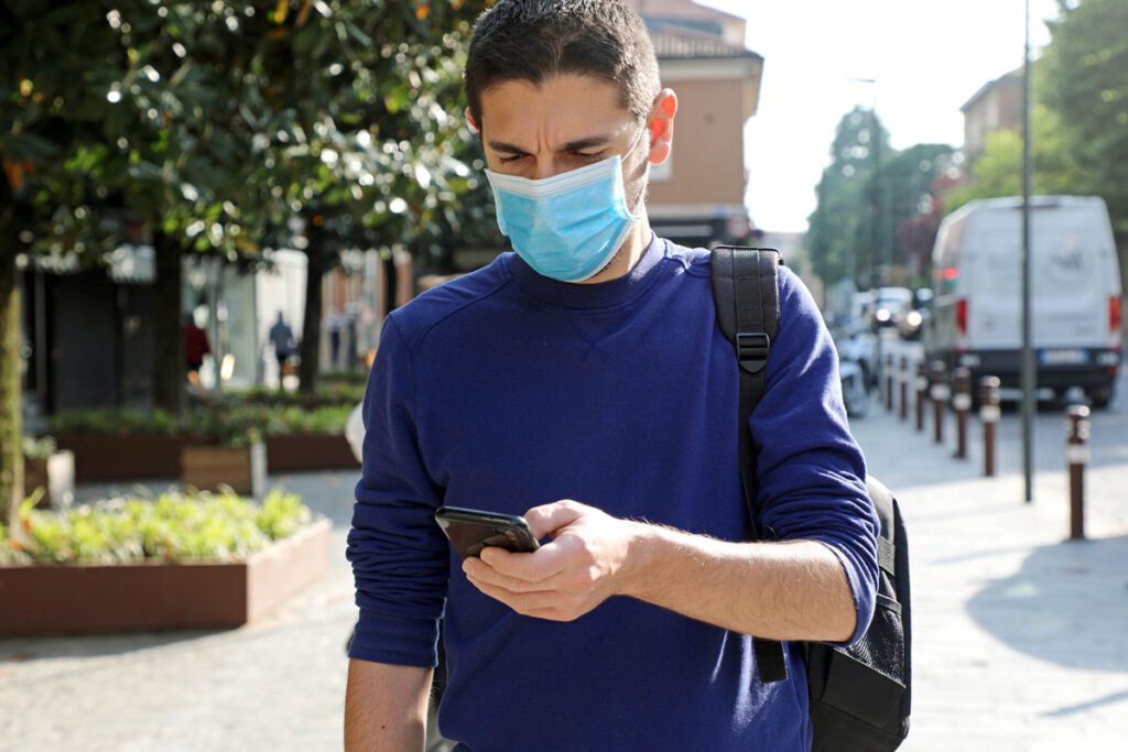 Worried Young Man Wearing Surgical Mask Using Smart Phone App in City Street to Aid Contact Tracing, representing the Insight Global data breach class action lawsuit settlement.