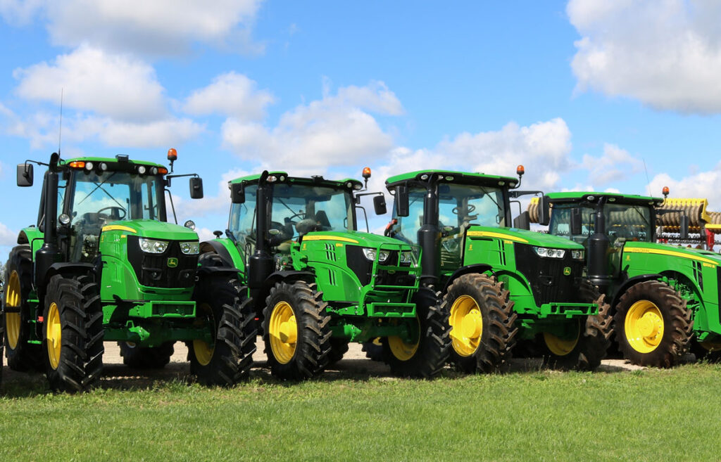 Row of green and yellow John Deere tractors for sale lined up on a field against a blue sky.
