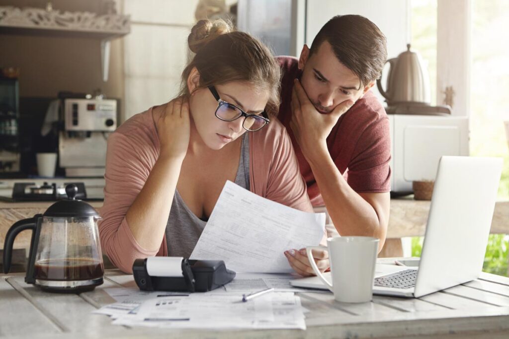 Female in glasses and brunette man studying paper form bank while managing domestic budget together in kitchen interior.