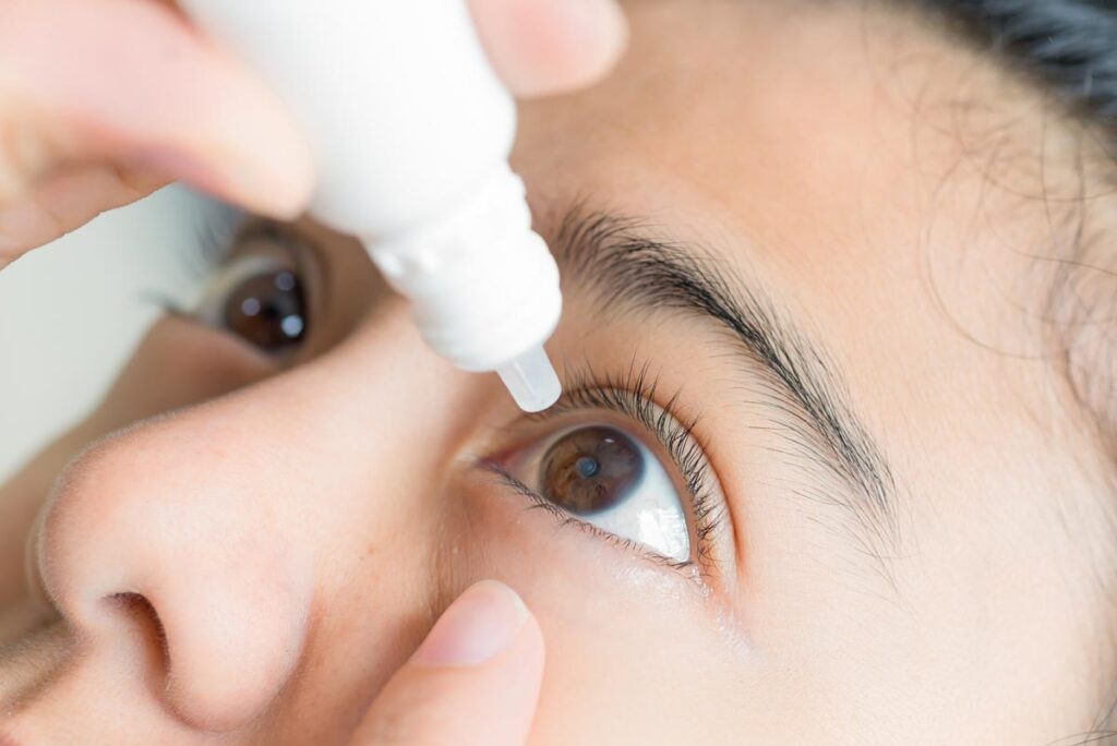 Close up view of young woman applying eye drop to left eye.
