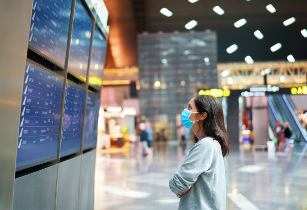 A woman at an airport looks at flight information board with a mask on, representing the Lufthansa class action lawsuit settlement.